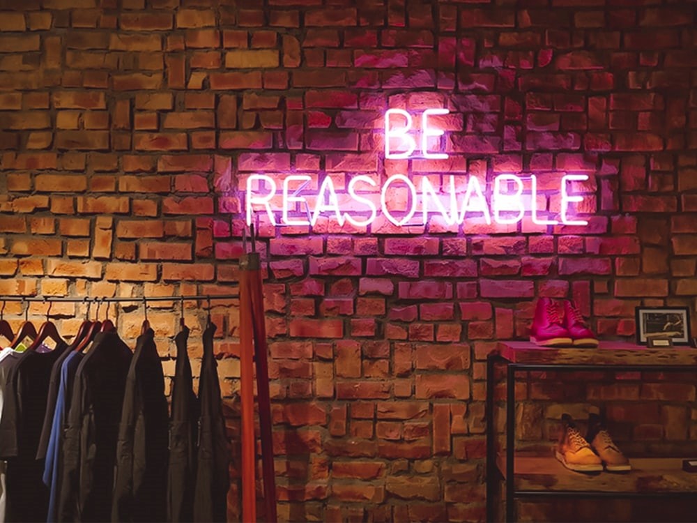 neon light that says be reasonable against brick wall